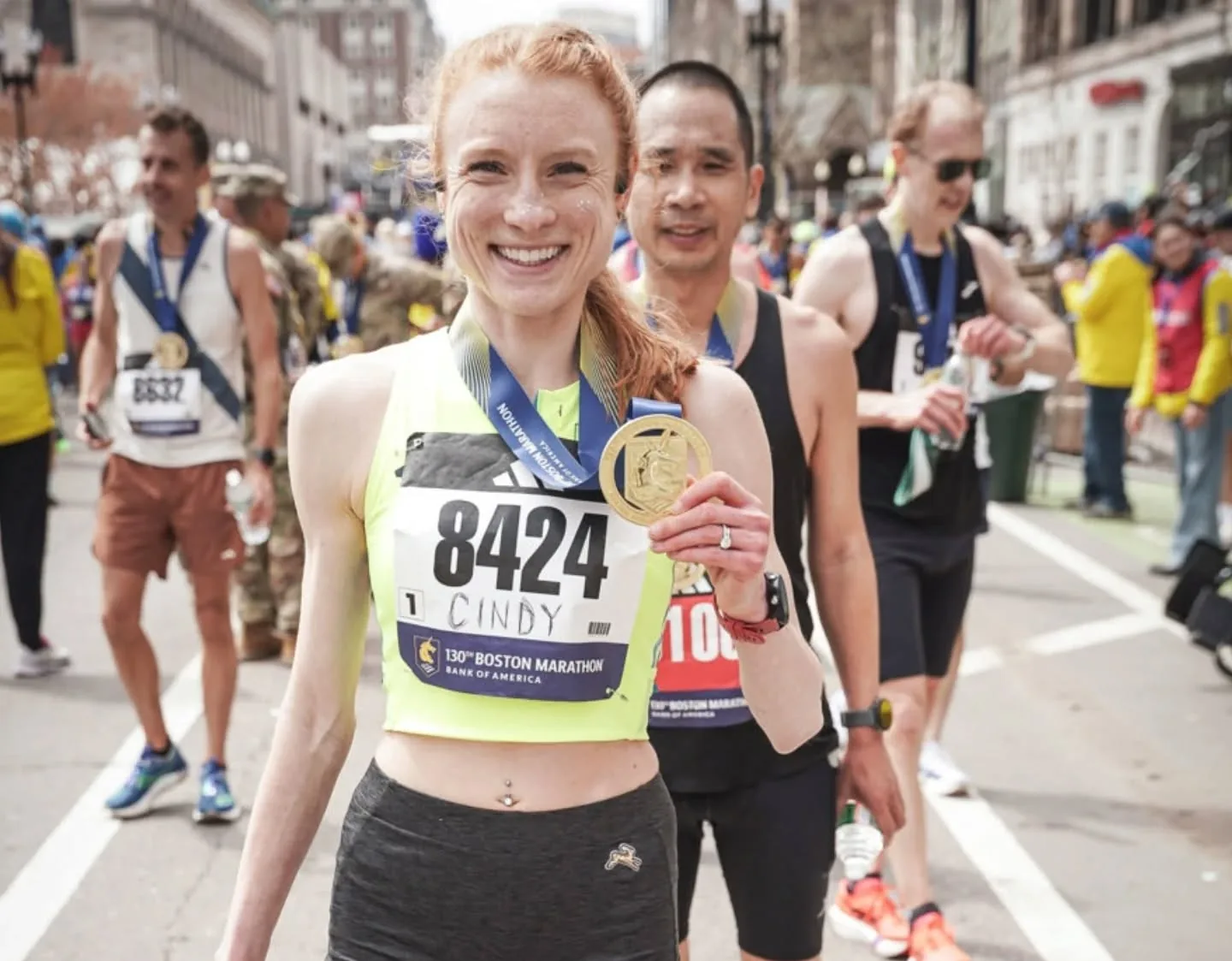 Cindy smiling and holding up her gold Boston Marathon finisher medal at the finish area